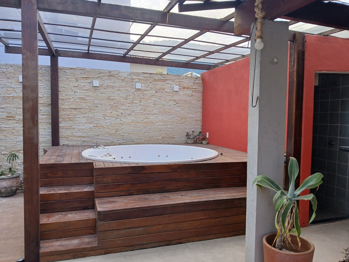 An outdoor area features a wooden platform with steps leading up to a round hot tub. The surrounding walls are adorned with textured stone, contrasting with a vibrant red wall. Natural light filters through a transparent roof, enhancing the space's inviting feel.
