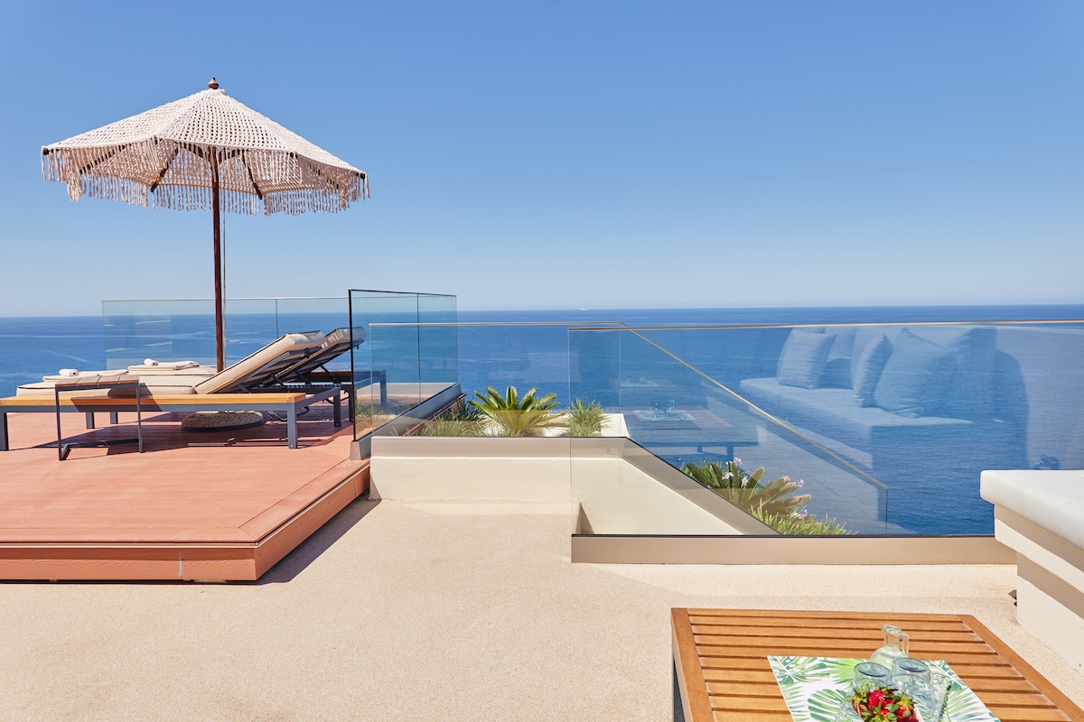 An outdoor terrace features a sun umbrella providing shade over a comfortable lounge area. Two sun loungers face the ocean, separated by a glass barrier that enhances the view. A wooden table holds a decorative arrangement, complementing the serene coastal landscape.