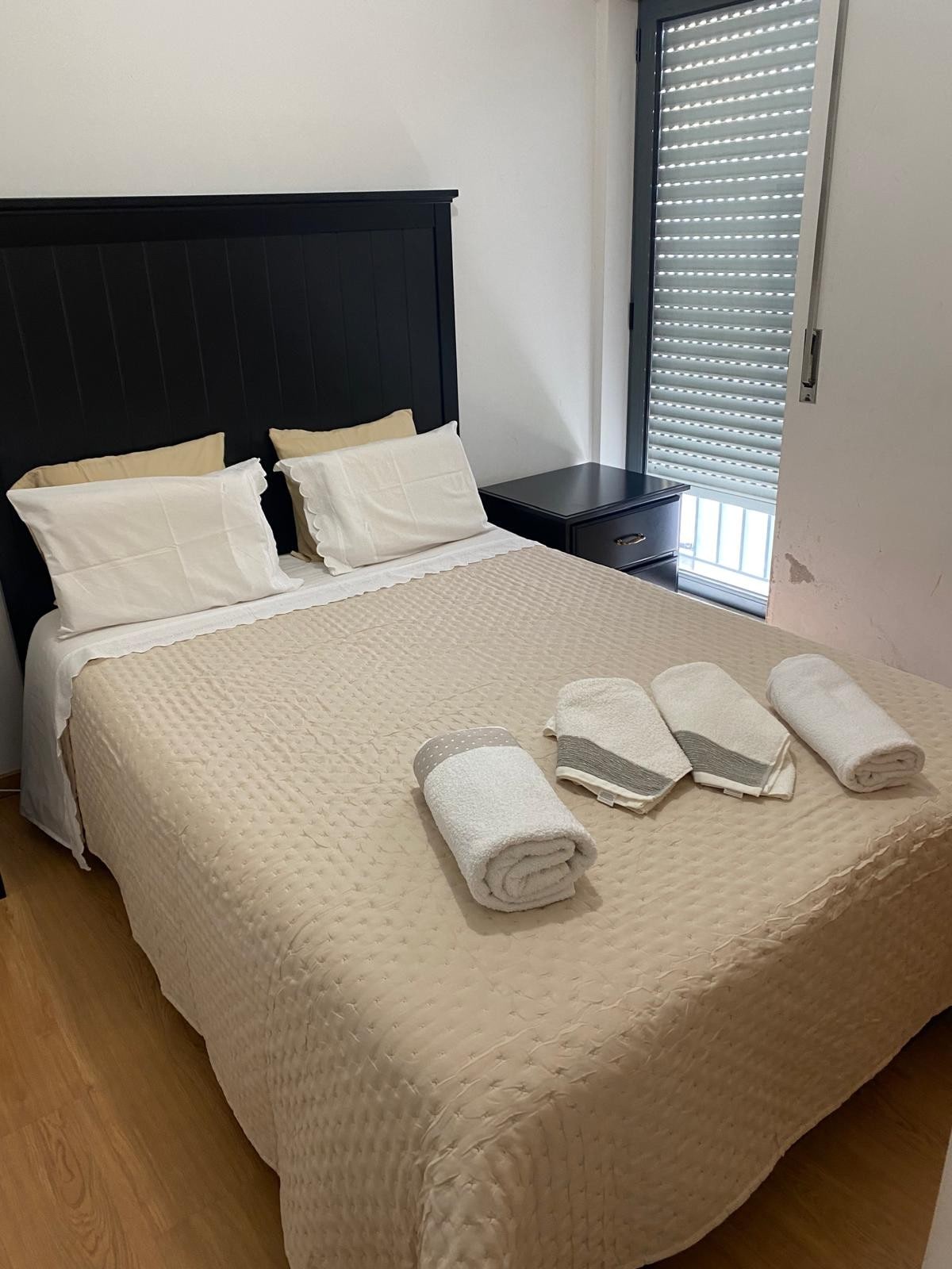 A cozy sleeping area features a spacious bed with a neutral quilted coverlet and two lightly cushioned pillows. Fresh towels are neatly arranged at the foot of the bed. Natural light filters in through a partially closed window with modern blinds, complementing the warm wooden flooring.