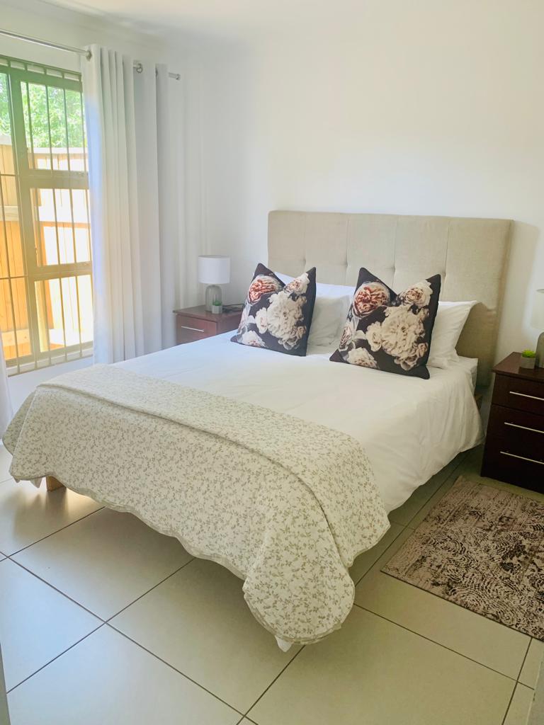 A well-appointed bedroom features a spacious bed adorned with decorative pillows. Soft light filters through sheer curtains, enhancing the calming atmosphere. Two nightstands sit on either side, each topped with a lamp, while a patterned rug complements the tiled floor.