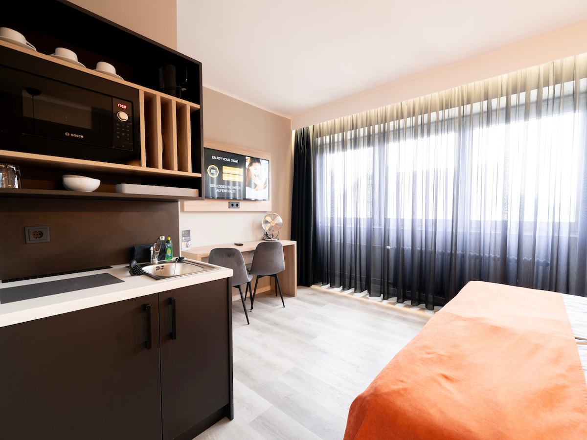 A comfortable studio apartment is showcased, featuring a kitchenette with a microwave and a small sink. Large windows dressed in sheer black curtains allow natural light to fill the room. A compact desk with two chairs and a 43" smart TV are visible, alongside a cozy bed with an orange throw.