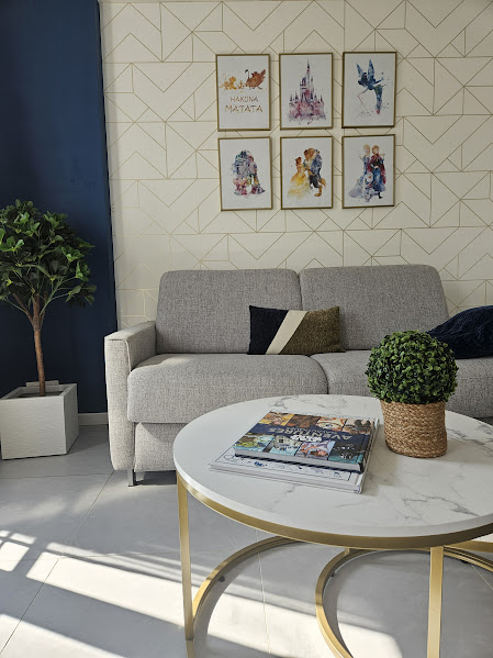 A light-filled living area showcases a gray sofa adorned with decorative cushions. A round marble coffee table with a gold base is centered in front, featuring magazines and a small potted plant. A green leafy plant is positioned in a white planter beside the sofa against a patterned wall.