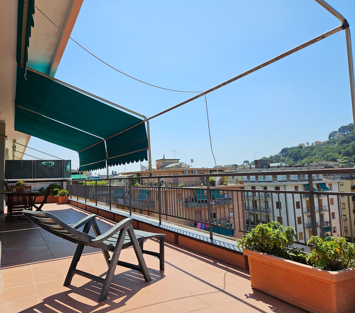 A spacious terrace features a sunbed and potted plants, with green awnings providing shade. The view includes surrounding buildings and a distant hillside, all under a clear blue sky.