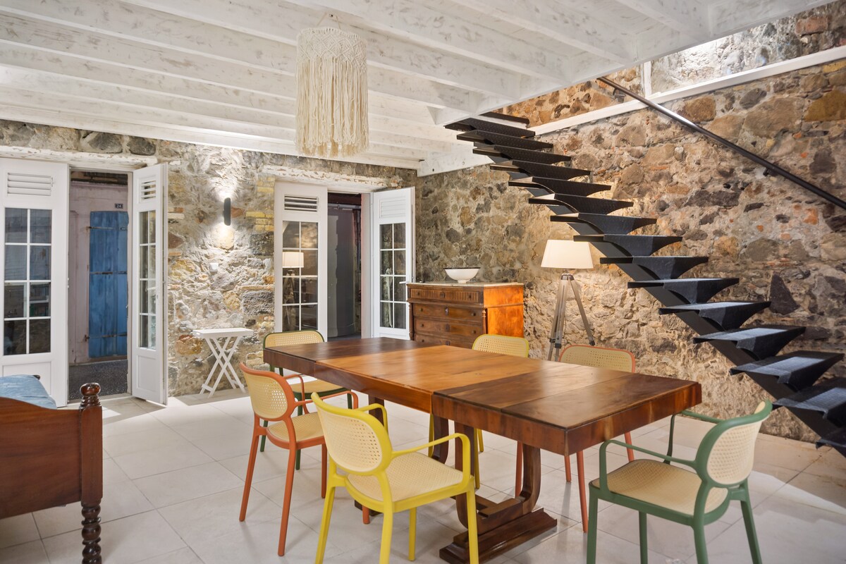 A spacious dining area features a large wooden table surrounded by colored chairs. The room showcases exposed stone walls and has large glass doors that open to the outside. A modern staircase leads to the upper levels, illuminated by a stylish light fixture.
