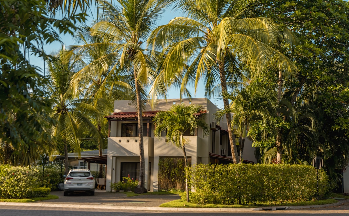 A tropical beach house is showcased amidst lush landscaping and tall palm trees. The two-story structure features a welcoming façade with large windows and a covered porch area. A white vehicle is parked in front, adding a sense of accessibility to the inviting surroundings.