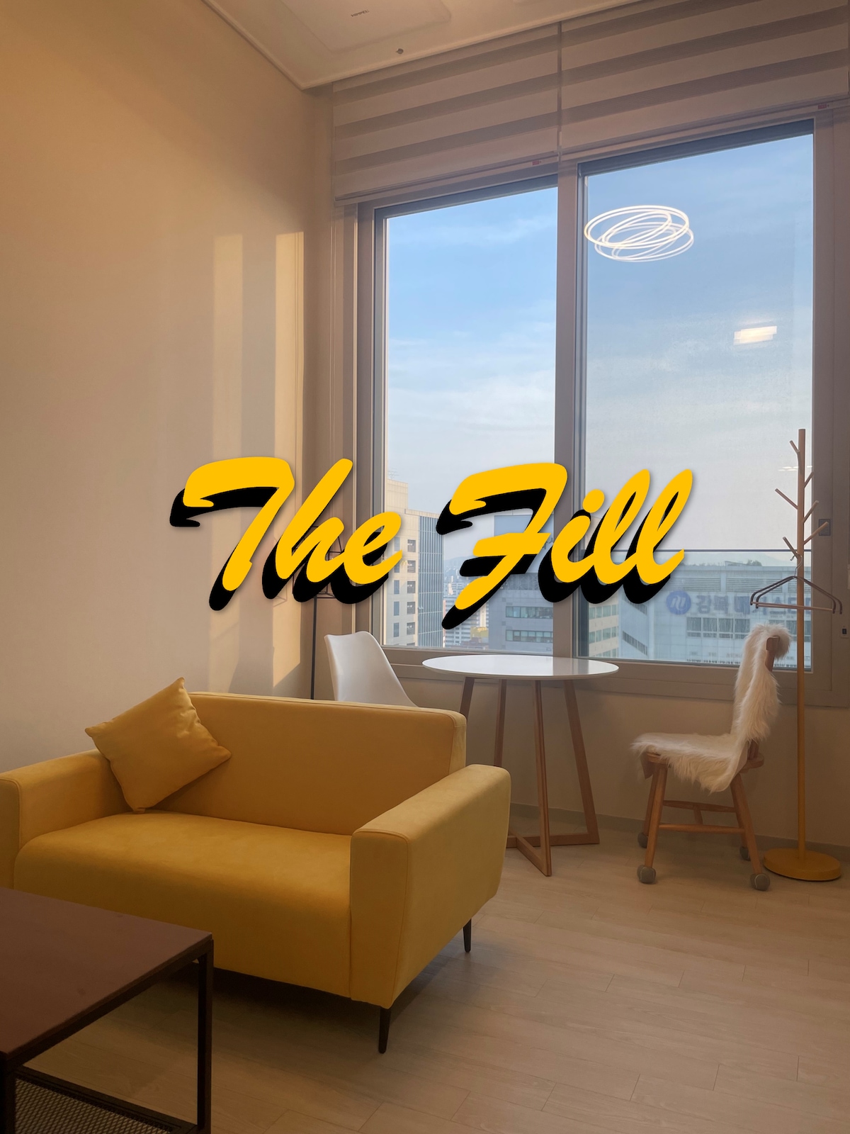 A bright living area is shown featuring a yellow sofa with a decorative pillow. A round dining table with two chairs is positioned by a large window, allowing natural light to fill the space. A unique coat rack adds a touch of functionality.