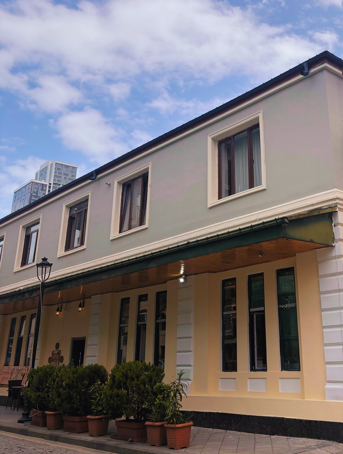 The exterior of the building is characterized by a light-colored façade, large windows, and a covered entrance with hanging lights. Potted greenery lines the base of the walls, and a contrasting darker floor adds depth to the welcoming space.