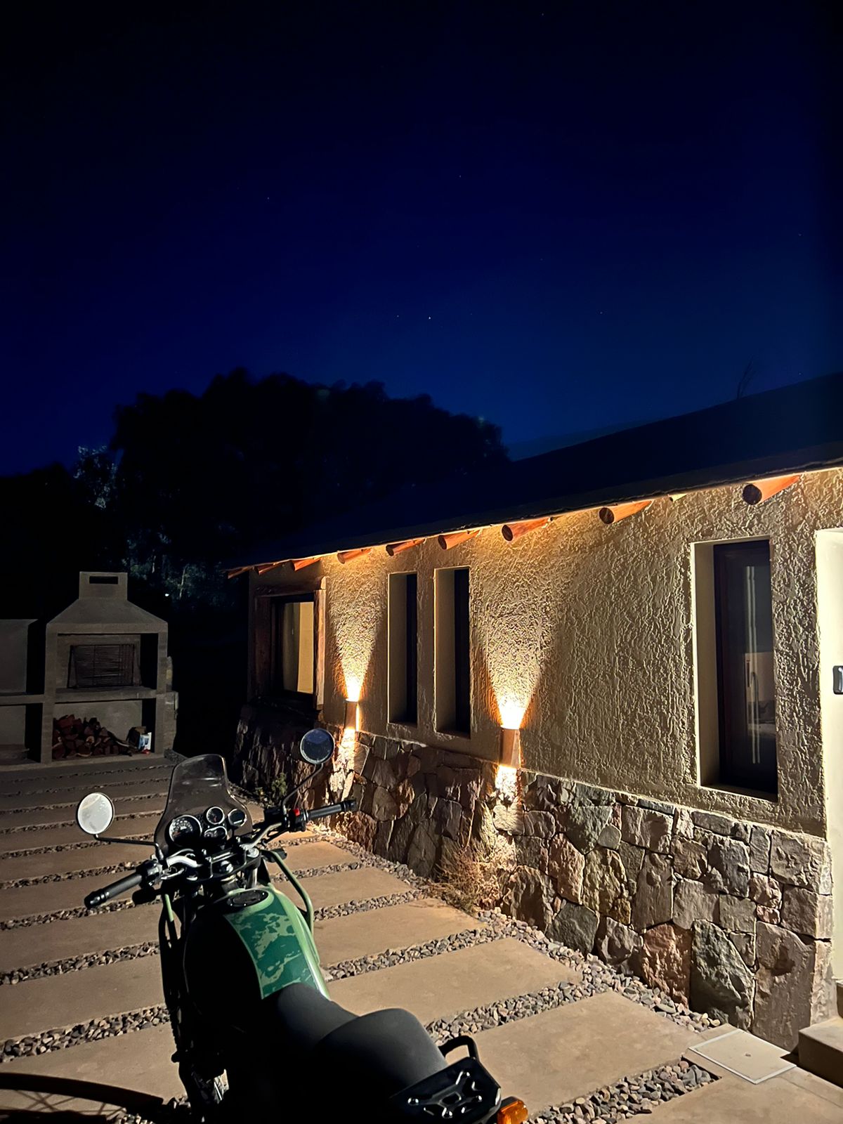 The exterior of a cabin is illuminated at night, highlighting textured stone and warm lighting. A motorcycle is parked nearby on a path lined with small stones, leading towards large windows that reflect the serene night sky.