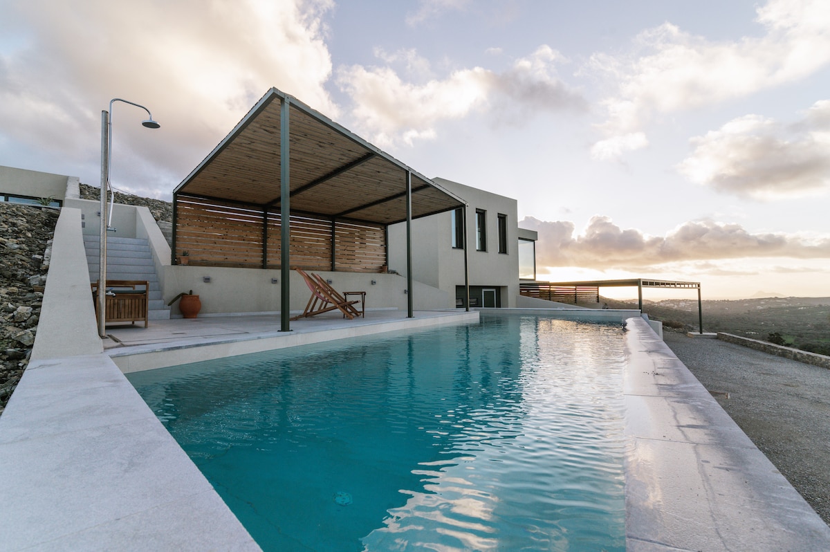 A serene pool area is highlighted by a modern structure with a wooden overhang. The pool's clear water reflects the sky's soft colors, creating a calming ambiance. Surrounding the pool, a minimalistic landscape enhances the peaceful setting with views of the hillside.