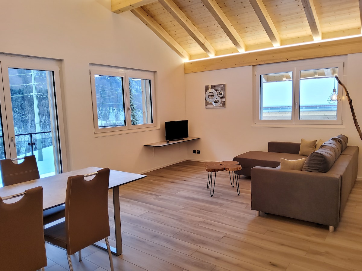 A modern living space features a comfortable sectional sofa facing a wall-mounted television. Large windows provide natural light, while a wooden dining table with four chairs sits nearby. The wooden ceiling adds an element of warmth to the open layout.
