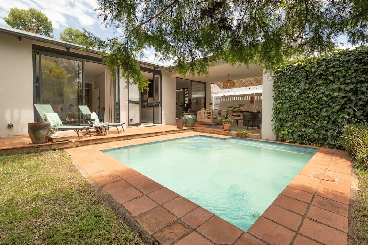 A serene outdoor space featuring a sparkling swimming pool framed by terracotta tiles. Sun loungers are positioned beside the pool, and lush greenery surrounds the area, providing a tranquil setting for relaxation and outdoor dining.