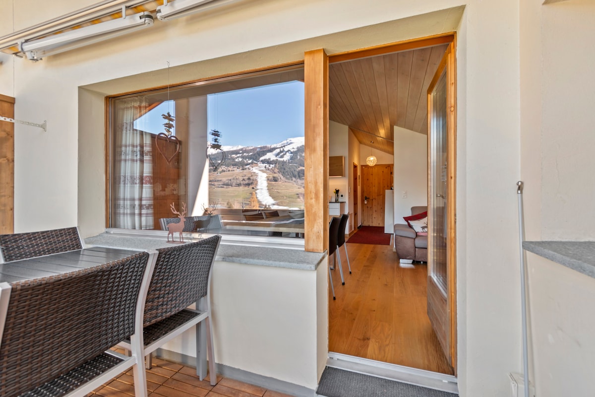 A large window frames a stunning view of the mountainous landscape. The balcony features a dining table with woven chairs, inviting outdoor meals. The warm, wooden interior is visible through the open door, enhancing the connection between indoor and outdoor spaces.