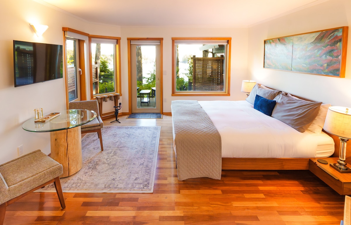 A spacious room features a king-sized bed with a soft grey blanket and two bedside lamps. A dining table made from natural wood stands beside a large window, offering views of the outdoor patio. A television is mounted on the wall opposite the bed.