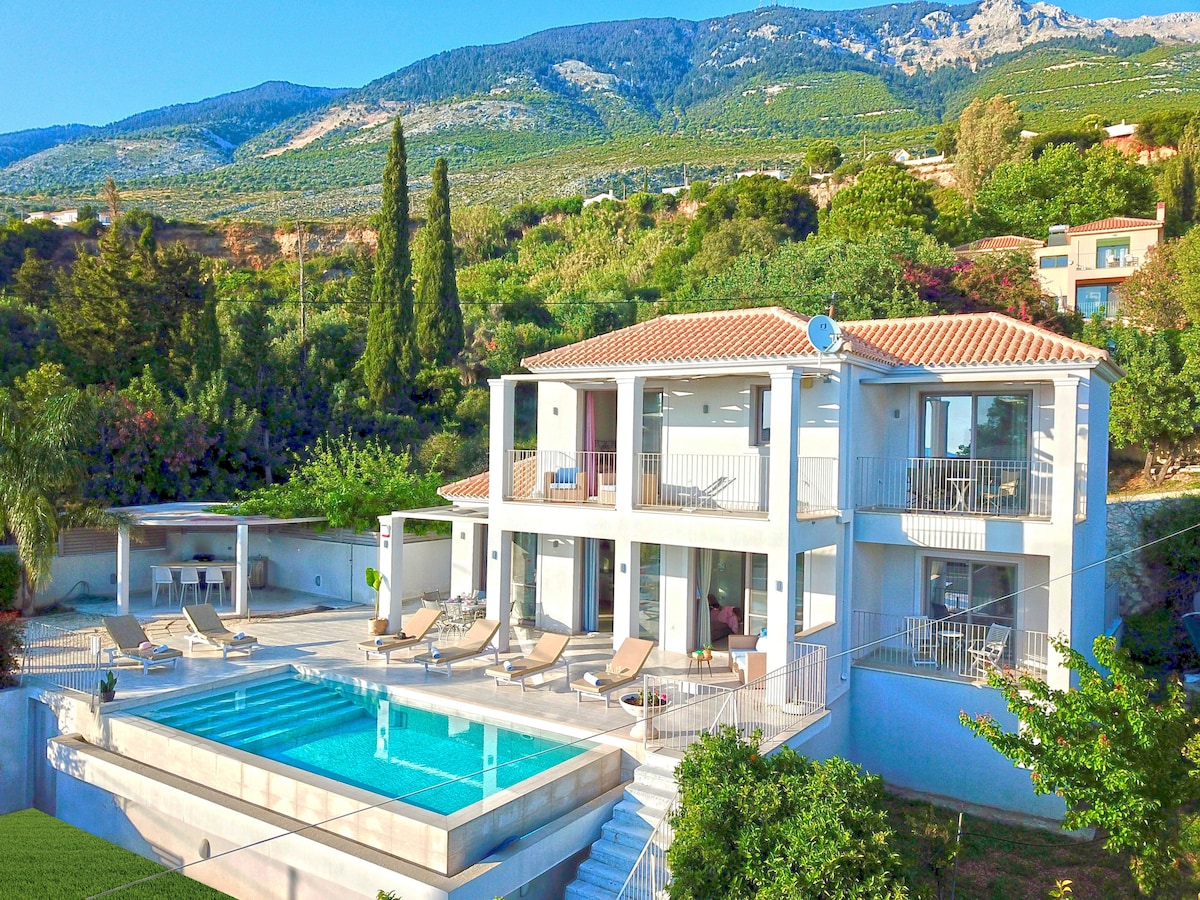 A spacious two-story villa is surrounded by lush greenery and mountains in the background. An inviting swimming pool is situated in front, accompanied by several sun loungers. Expansive glass doors provide a view into the interior, enhancing the connection with the outdoor setting.