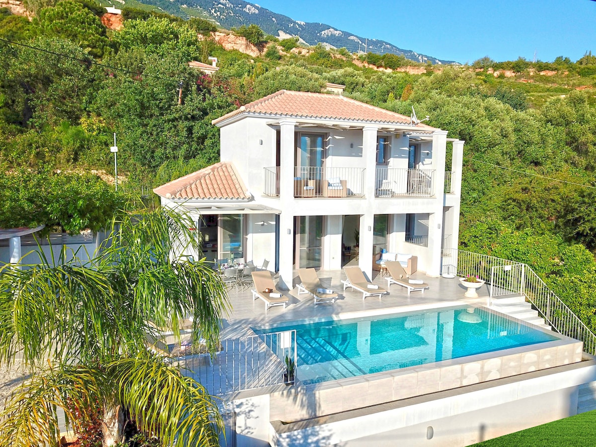 A modern two-story villa is surrounded by greenery, featuring a spacious pool area with sun loungers. A covered terrace overlooks the pool, which is bordered by a sleek coping. The light-colored exterior contrasts with the vibrant natural landscape and distant mountains.