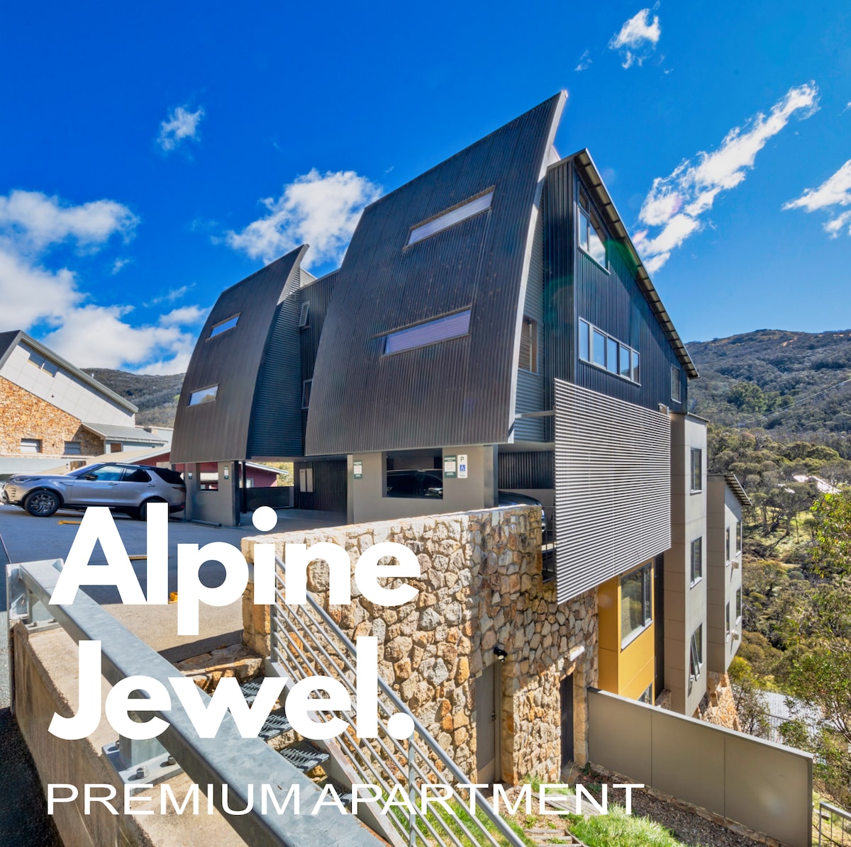 The exterior of the apartment showcases a modern architectural design, featuring angled roofs and a mix of natural stone and wood materials. The sunlight highlights the unique building structure against a backdrop of mountains and blue skies.