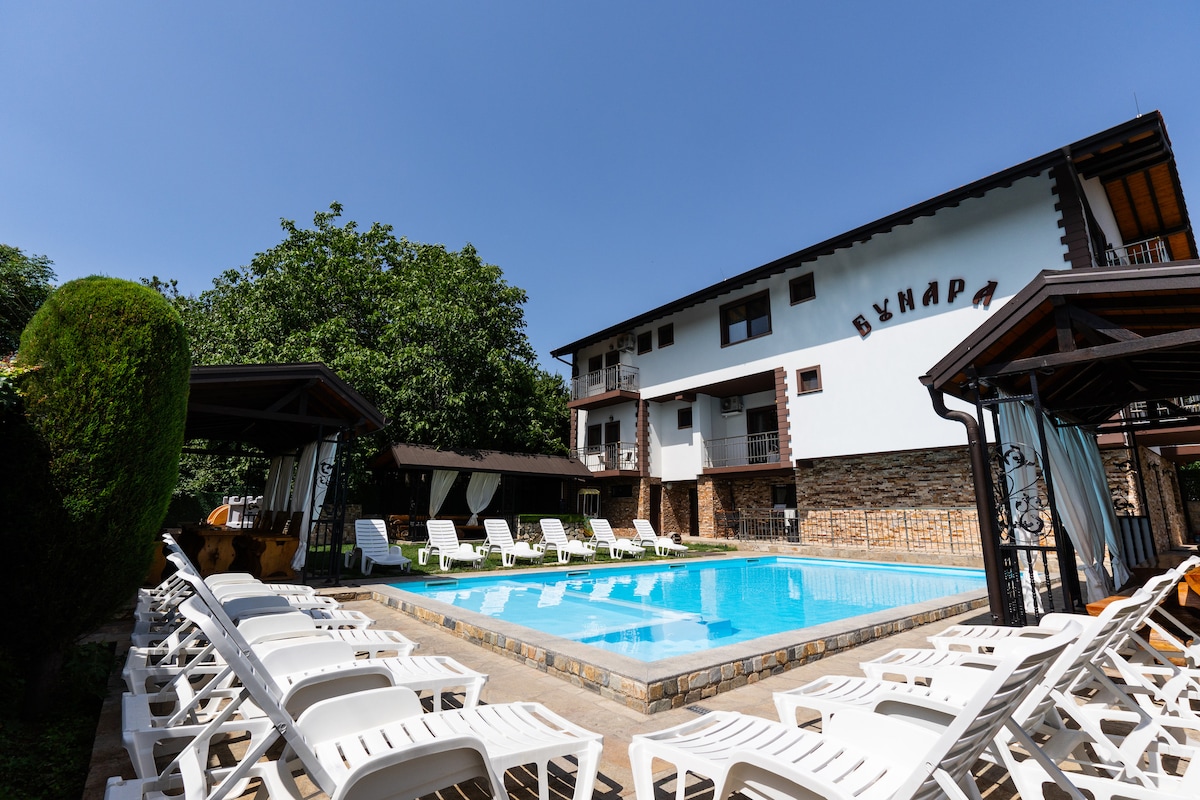 The outdoor area features a refreshing swimming pool surrounded by comfortable lounge chairs. A well-maintained garden and shaded cabanas complement the space, providing an inviting atmosphere for relaxation. The multi-story building adds a functional backdrop to the sunny setting.