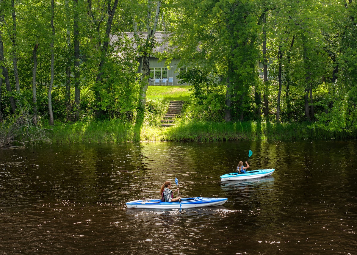Two kayakers are paddling on the calm river, surrounded by lush green trees. A cottage with large windows is visible on the riverbank, backed by a staircase leading to the water. The scene reflects a peaceful outdoor environment.