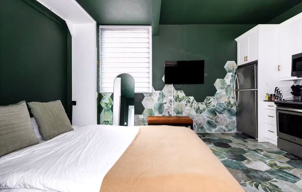 A spacious studio apartment features a queen-sized Murphy bed with a neutral-colored throw blanket. Green walls adorned with hexagonal patterns complement the sleek white cabinets and modern appliances in the kitchenette. Natural light is invited through the window, illuminating the vibrant floor design.