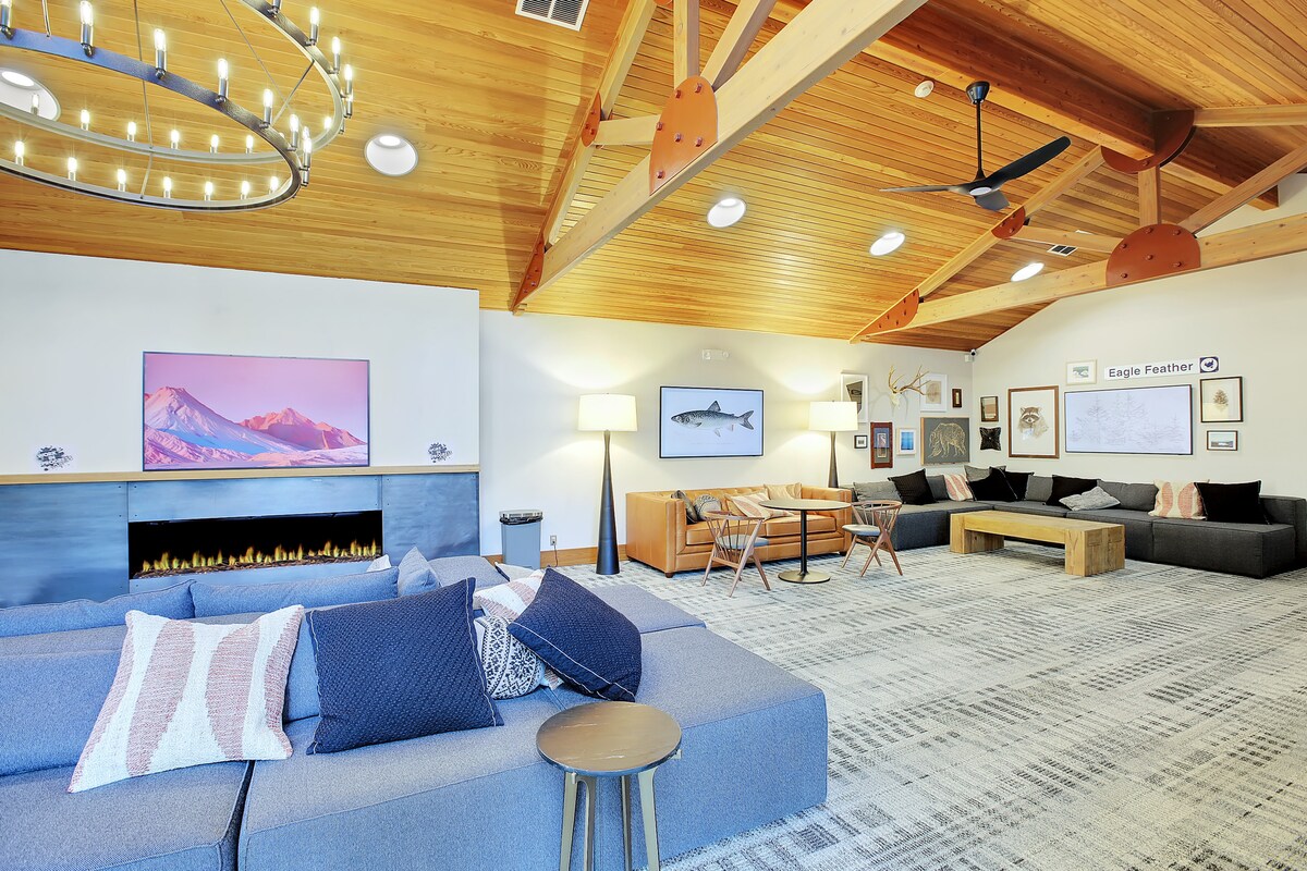 A spacious common area features high ceilings with exposed wooden beams and ambient lighting from a circular chandelier. Comfortable seating is arranged around a modern fireplace, with a large television mounted on the wall. The decor includes framed art and a unique blend of furnishings.