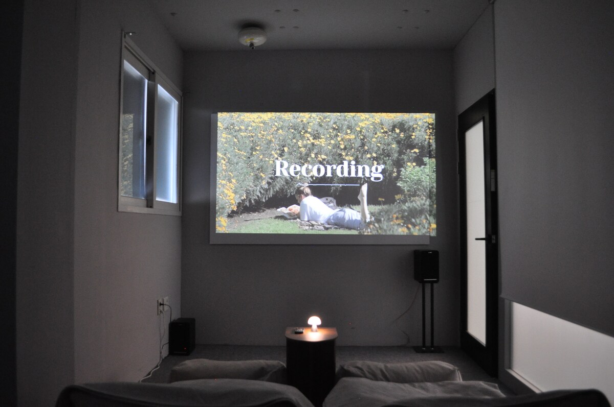 A beam projector displays a recording on a wall in a softly lit room. Comfortable seating arrangements are visible, accompanied by a side table with a lamp. Large windows allow natural light, while a speaker stands nearby, contributing to a cozy entertainment environment.