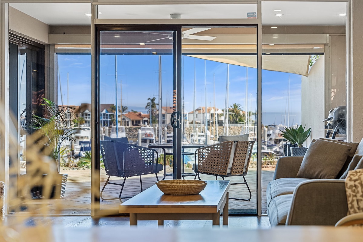 Large glass doors provide a clear view of the marina, where boats are docked under a clear blue sky. A comfortable seating area with woven chairs and a coffee table creates an inviting space. Bright natural light flows into the open living area.