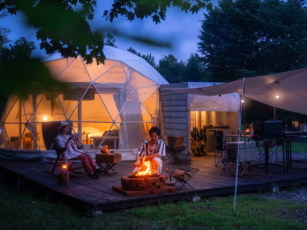 A glamping dome is illuminated from within, contrasted against the evening sky. Two individuals are seated by a fire pit on the wooden deck, creating a cozy atmosphere. The exterior features a spacious deck area with seating arrangements and cooking equipment under a large canopy.