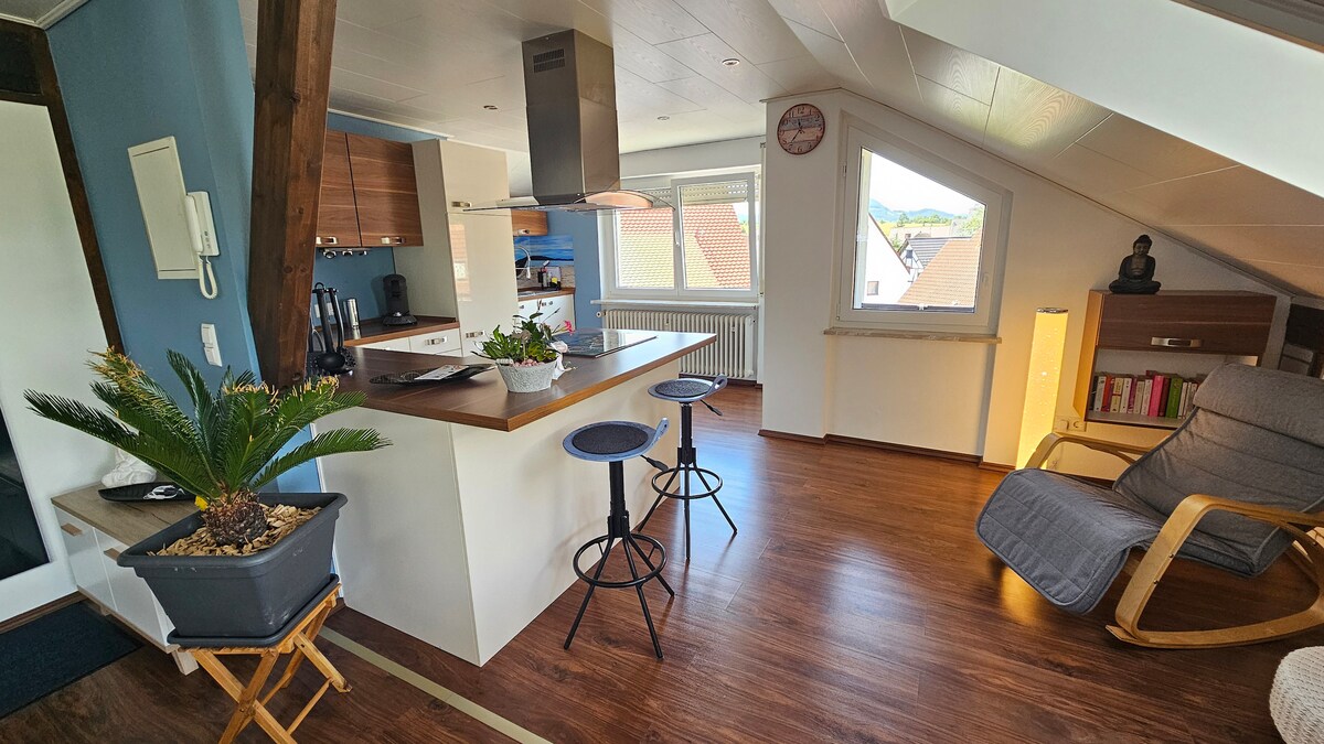 A modern open-plan kitchen and living area are presented, featuring sleek cabinetry and a wooden counter with two bar stools. A comfortable rocking chair is positioned nearby. Natural light fills the space through a large window, with a decorative plant adding a touch of greenery.