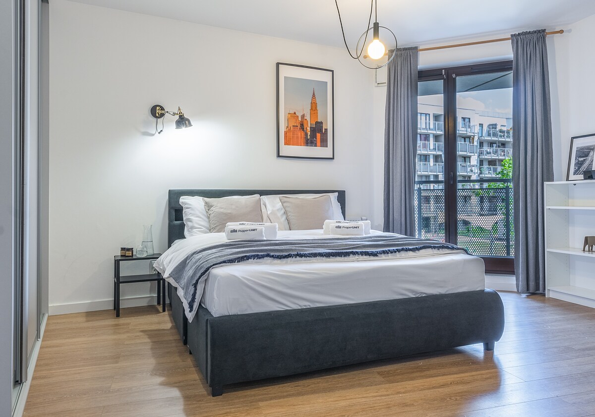 A spacious sleeping area features a comfortable bed dressed in soft linens. A framed cityscape hangs above the bed, while natural light filters through sliding glass doors that lead to a balcony. A stylish lamp and side table provide functional decor.