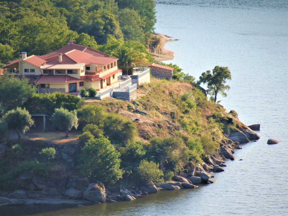 A spacious building with a red-tiled roof is nestled on a river peninsula, surrounded by greenery. The structure features multiple levels and balconies overlooking the water. Rocky shorelines and trees provide a natural border along the riverbank.