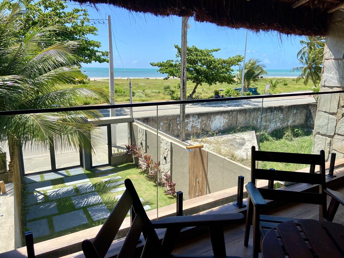 A view from a balcony overlooks a green expanse leading to the ocean. Bright blue skies and palm trees frame the scene, while a pathway with stone tiles and plants can be seen below. Outdoor seating is visible on the wooden deck.