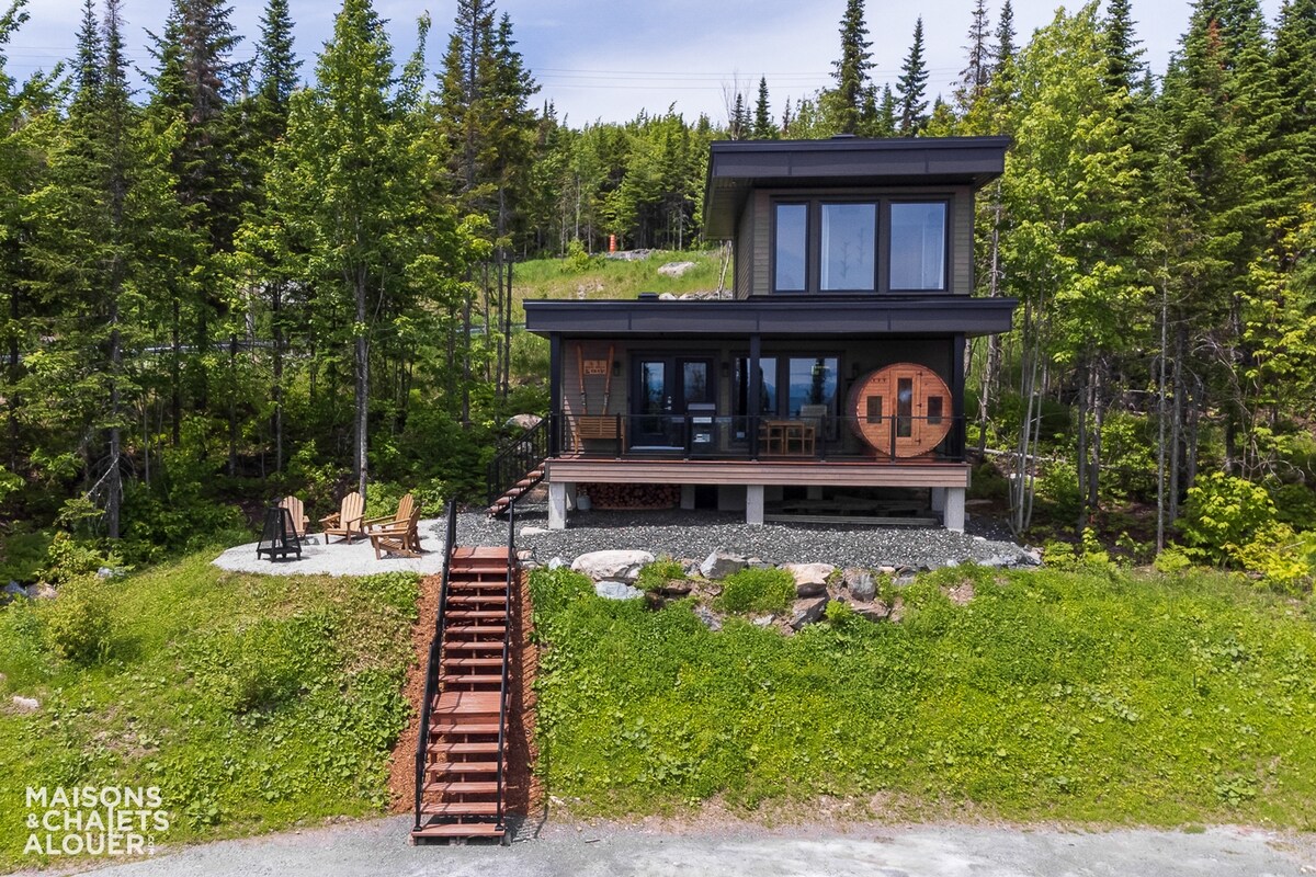 The chalet is framed by a lush natural landscape, featuring large windows that offer views of the surroundings. A wooden staircase leads to the entrance, and a seating area with chairs is positioned on the patio, providing a comfortable outdoor space.