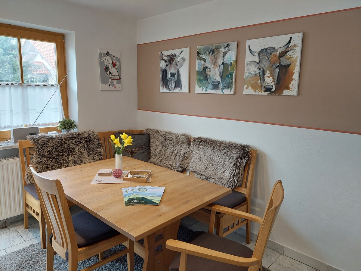 A dining area features a wooden table surrounded by a cozy corner bench and chairs. Four artistic animal portraits are displayed on the wall. A vase with fresh flowers is positioned on the table, with magazines laid out for easy access. Natural light enters from the window.
