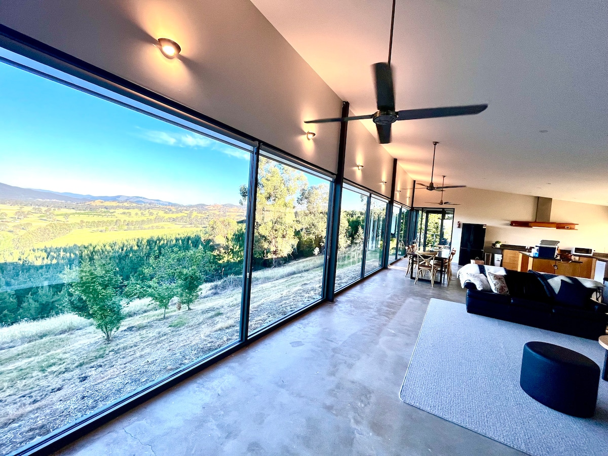 A spacious living area features expansive glass walls, providing unobstructed views of the surrounding landscape. Soft lighting enhances the open space, while modern ceiling fans add comfort. A dining table and a comfortable seating area with a dark couch are included, creating a welcoming atmosphere.