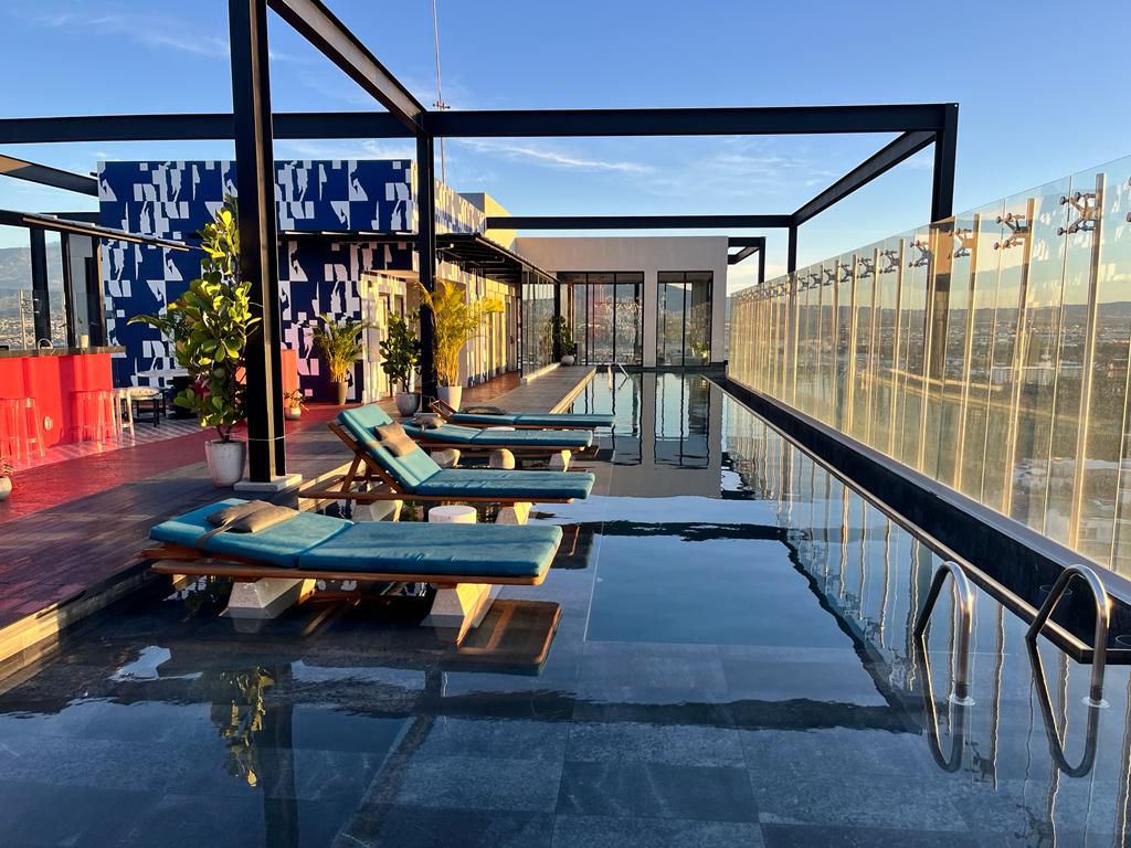 A rooftop pool area is highlighted, featuring lounge chairs positioned beside a sleek swimming pool. Surrounding the space, contemporary structures and vibrant decor are visible, with panoramic views of the skyline. Glass panels provide an airy feel while ensuring safety.