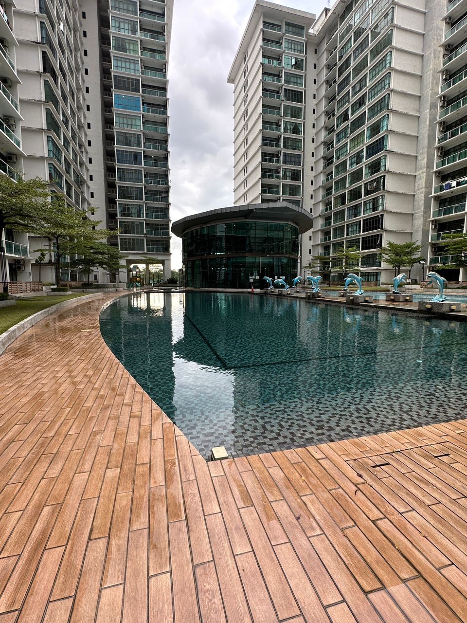 A serene pool area is framed by modern residential buildings on either side. The clear water reflects the sky and surrounding greenery, while wooden decking leads to inviting lounge chairs near the pool. Sculptures are positioned along the water's edge, enhancing the tranquil atmosphere.