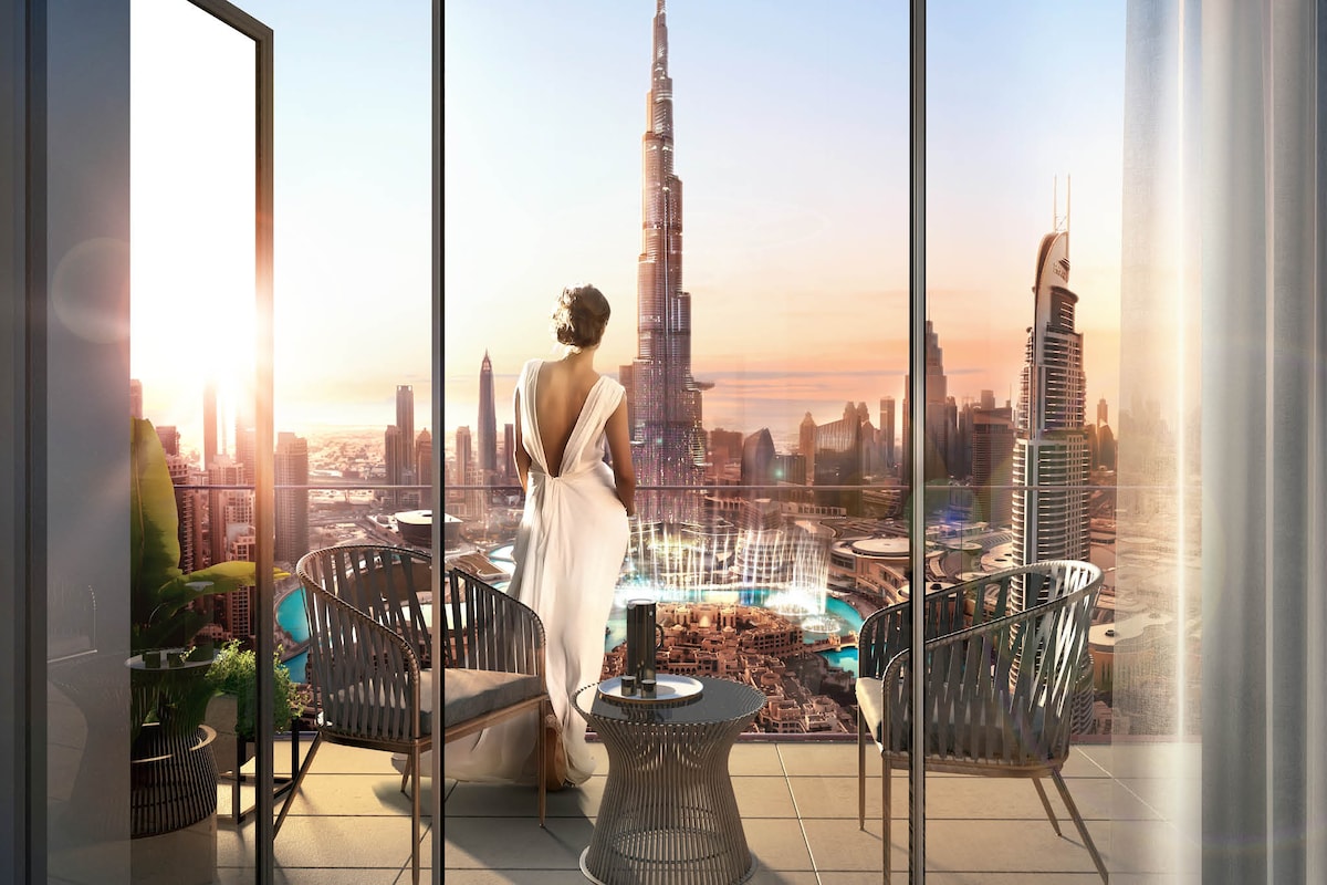 A spacious balcony offers stunning views of the Burj Khalifa and Dubai Fountain. Stylish outdoor seating includes two modern chairs and a small table. The scene is enhanced by warm sunlight, casting a soft glow over the surroundings, creating a serene ambiance.