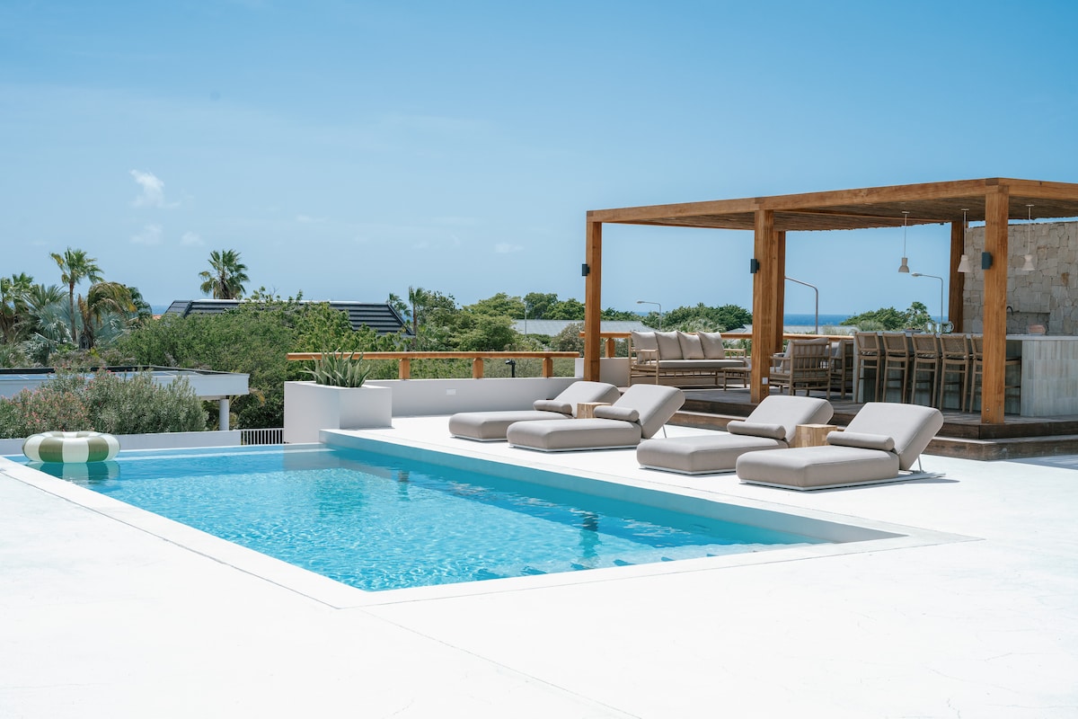 An infinity pool is featured, complemented by several lounge chairs in soft tones. A wooden structure nearby offers a shaded seating area with an elongated dining table and overhead lighting, enhancing the outdoor ambiance. Lush greenery and a clear sky serve as the backdrop.