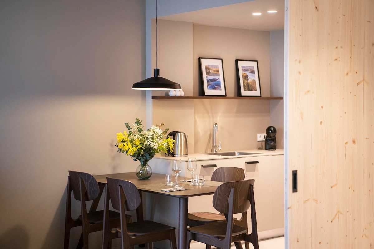 A dining area features a wooden table surrounded by four chairs. A floral arrangement is placed at the center, complemented by two glasses and cutlery. Above, a pendant light hangs, illuminating the space. The kitchenette is visible in the background, showcasing cabinetry and essential appliances.