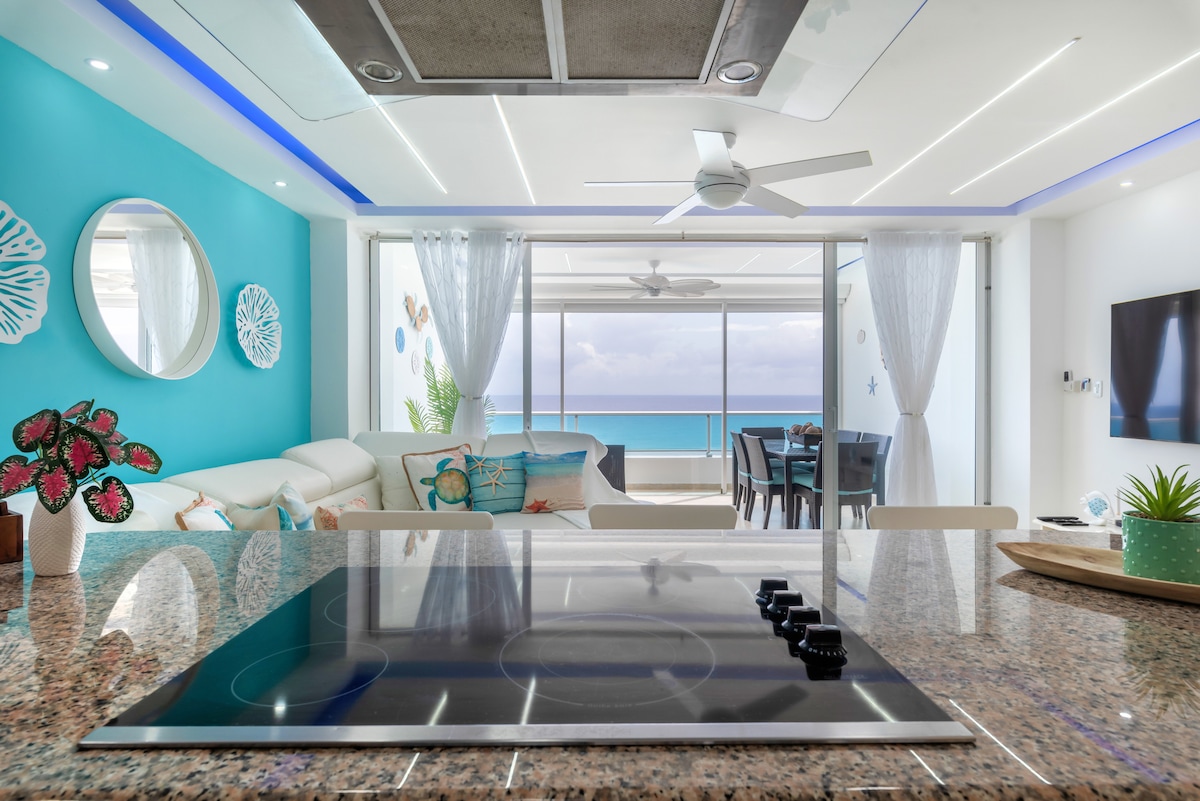 A modern kitchen is highlighted by a sleek countertop and an electric stove. The vibrant blue wall contrasts with the bright white decor. Clear views of the balcony and beach can be seen through large glass doors, enhancing a sense of openness within the space.