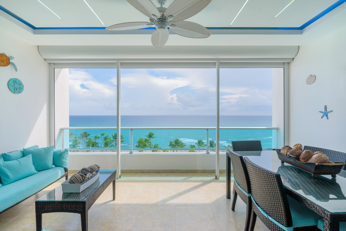 A bright balcony space is equipped with a dining table and couches, offering views of the ocean and palm trees. Natural light floods the area through expansive glass doors, complemented by a ceiling fan and decorative coastal elements on the walls.