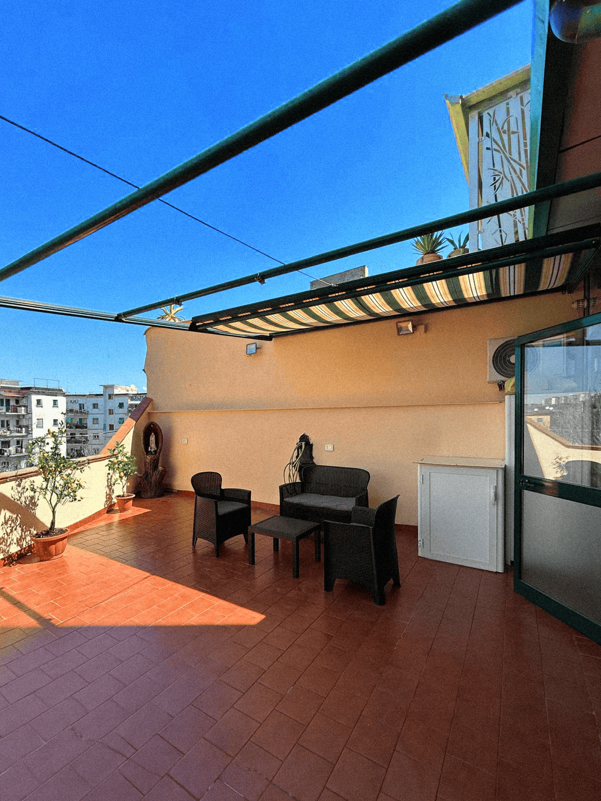 An outdoor terrace is shown, featuring a set of black chairs and a small table. Potted plants are placed around the area. A clear blue sky is visible above, with a glimpse of neighboring buildings in the background. A small storage unit is positioned against the wall.