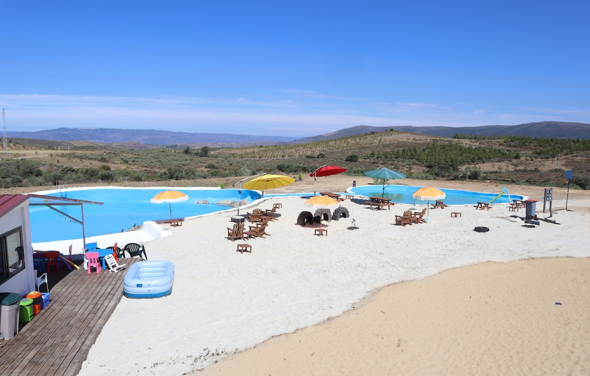 A spacious outdoor area features a large swimming pool surrounded by sandy beach-like terrain. Colorful umbrellas provide shade over picnic tables and lounge chairs. Scenic hills form the backdrop, with clear blue skies above, creating an inviting space for relaxation and leisure.