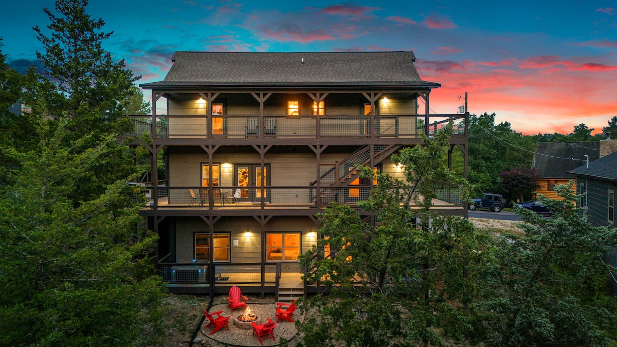 The cabin is showcased from an aerial view, revealing three levels with multiple balconies and outdoor seating. Red chairs surround a stone fire pit in the foreground, inviting gatherings under the sky, while lush greenery frames the scene amidst a colorful sunset.