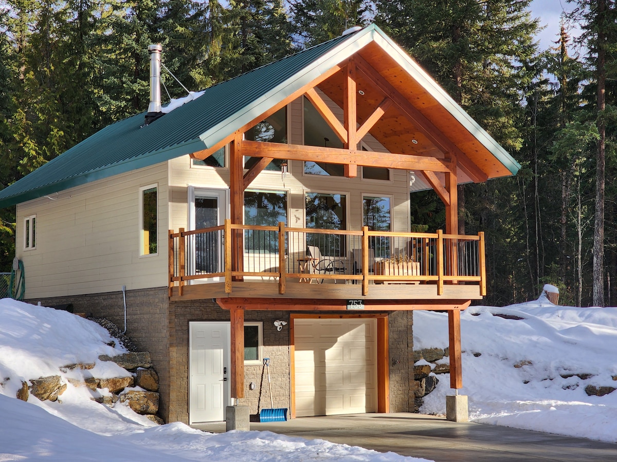 A charming cabin features a wooden exterior with a sloping green roof and exposed beams. A covered deck provides seating, while large windows allow ample natural light into the living area. A garage is located beneath the cabin, surrounded by a snowy landscape.
