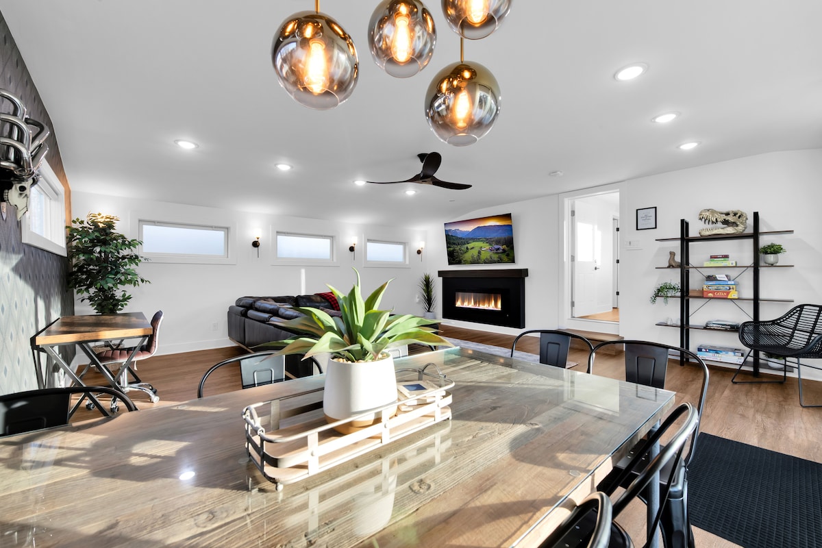 A modern living area is highlighted by a spacious wooden dining table with black chairs. Decorative light fixtures hang overhead, illuminating the room. A large sectional sofa is positioned toward a wall-mounted TV, and a cozy electric fireplace is visible, enhancing the inviting atmosphere.