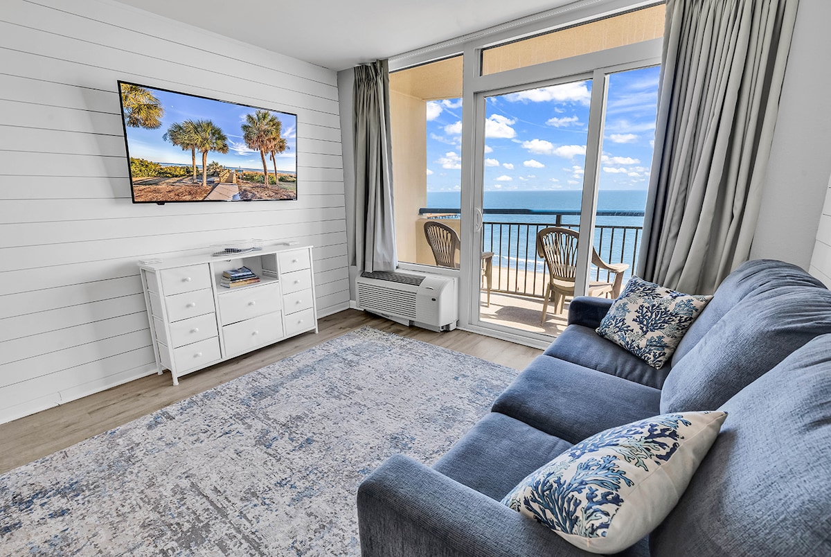 A bright and airy living room features a comfortable blue sofa paired with decorative pillows. A flat-screen TV is mounted on the wall, providing entertainment options. Large sliding glass doors reveal a view of the balcony, with palm trees and the ocean visible beyond.
