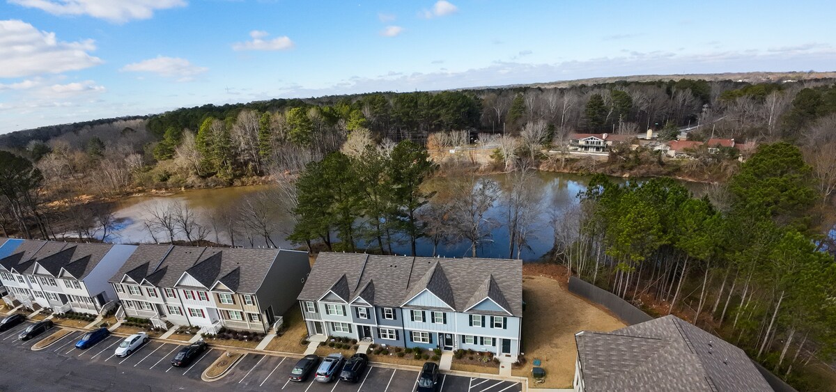 An aerial view captures a row of townhomes lined along a parking lot, with lush trees and a lake in the background. The tranquil water reflects the blue sky and scattered clouds, providing a serene natural setting.