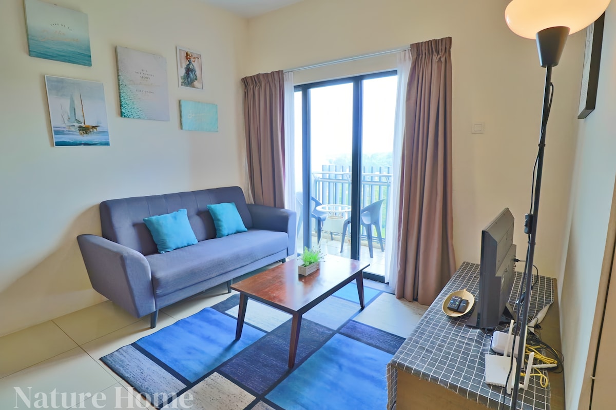 A comfortable living area features a gray sofa facing a flat-screen television. A glass door leads to a balcony, framed by light curtains. A coffee table sits on a patterned rug, with artwork displayed on the walls, adding visual interest to the space.