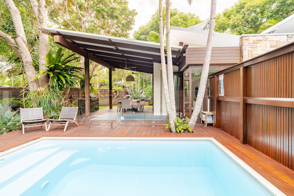 A private heated magnesium pool is set amidst a spacious wooden deck, featuring tropical plants and trees that provide lush greenery. The outdoor dining area is covered, allowing for comfortable gatherings, while two lounge chairs are positioned for relaxation under the sun.
